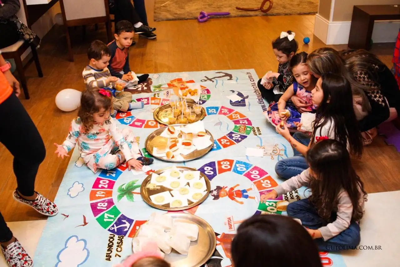 Crianças ao redor de um tabuleiro gigante com pratinhos de doces no chão de uma casa