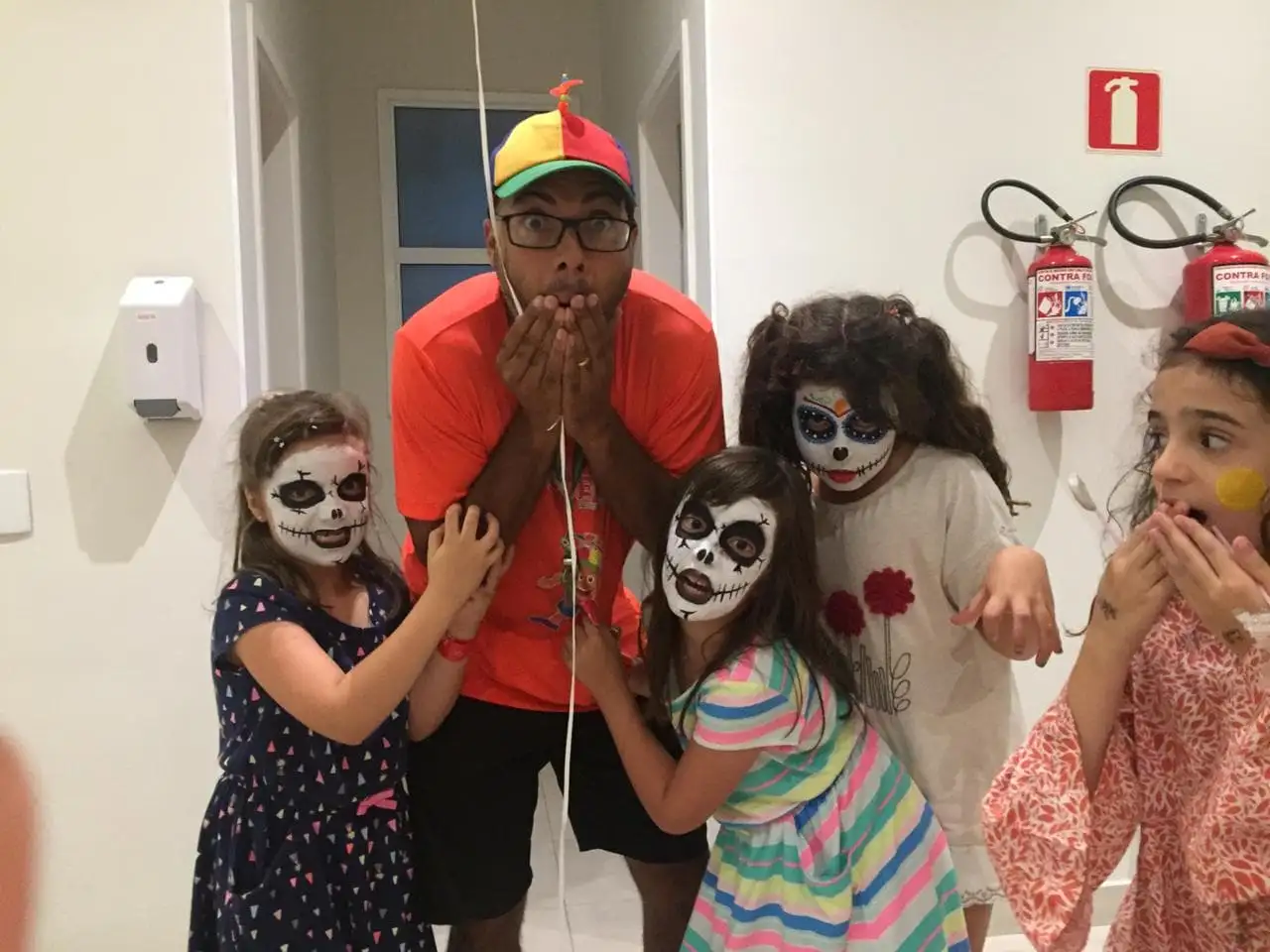 Tio Buchecha, de boné de hélice, faz cara de susto ao lado de crianças com o rosto pintado de caveira