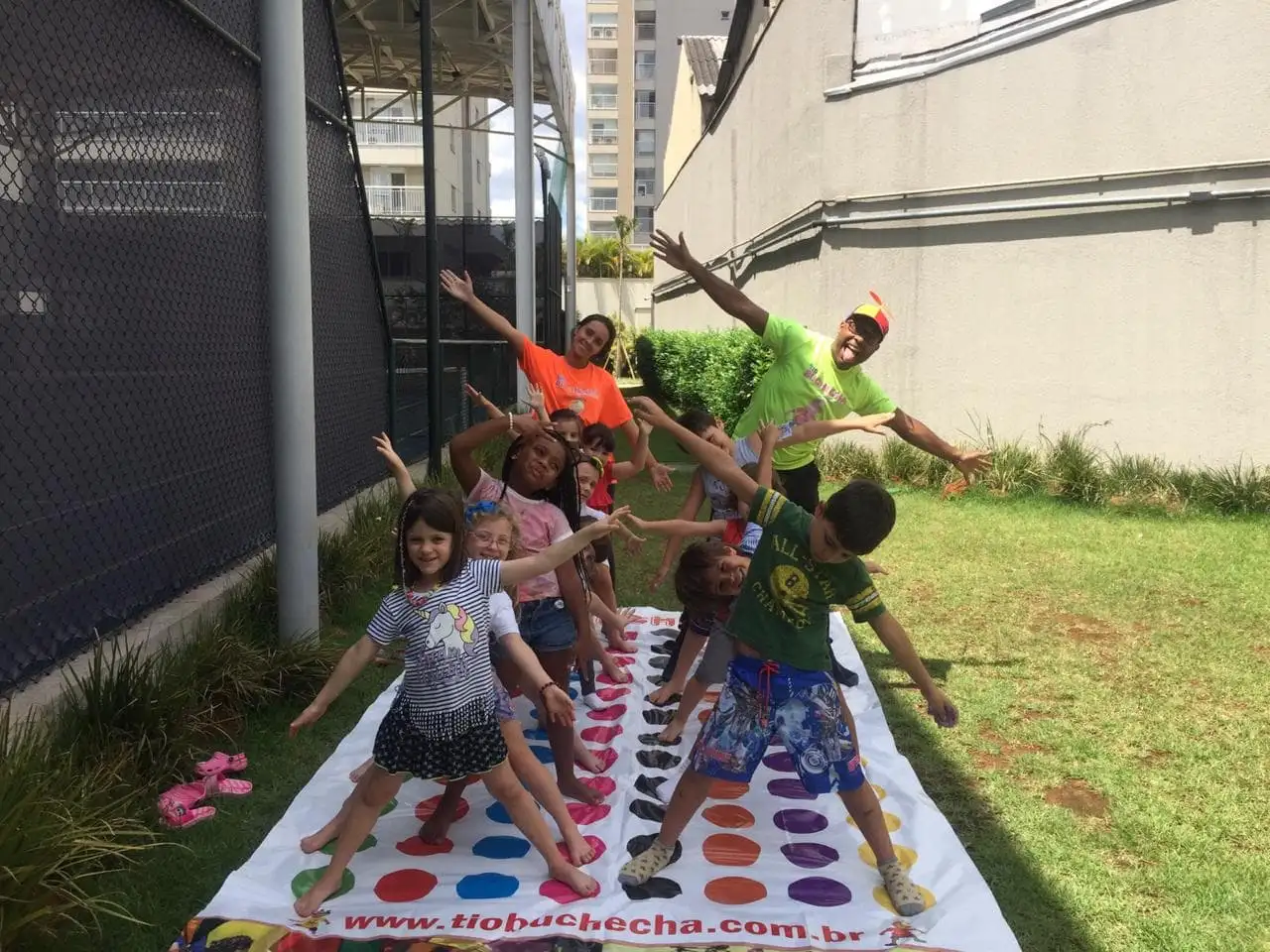 Crianças e recreadores abrem os braços em pose de estrela sobre um tapete de Twister no jardim