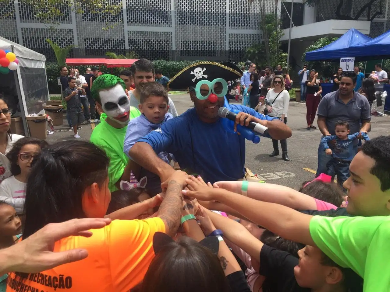 Crianças e recreadores fantasiados juntam as mãos no centro da roda num evento ao ar livre