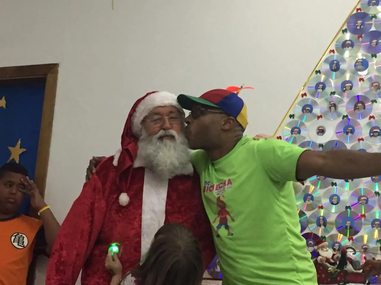 Tio Buchecha, de boné de hélice, dá um beijo na bochecha do Papai Noel